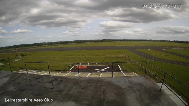 An outdoor view from a webcam at Leicesterhire Aero Club showing a runway and grassy airfield under a partly cloudy sky. In the foreground, there is a small helipad with a white helicopter symbol painted on it, enclosed by a black railing, and a windsock on a pole to the left. The background features a wide stretch of runway and open fields extending to the horizon. The timestamp in the top right corner indicates it was taken on April 6, 2025, at 13:00:57.