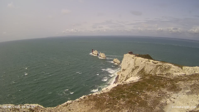 A view from a webcam showing white chalk cliffs extending into the ocean under a cloudy sky. There is a lighthouse on a small rock formation offshore, and a building on top of the cliffs. The sea is slightly choppy with white waves near the shore. The photo has a timestamp of "04/06/2023 14:00:07" in the bottom left corner.