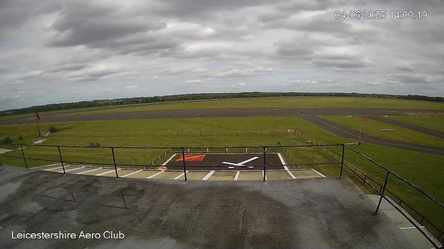 A wide view of an airport runway and surrounding grassy fields under a cloudy sky. In the foreground, there is a helipad marked with a large white "H" on a red background, with white lines and safety markings. The scene appears to be taken from an elevated position, likely from a webcam mounted on a building or control tower at the Leicester Aero Club, as indicated by the text at the bottom-left corner of the image. The timestamp in the top right corner reads "04-06-2025 14:00:19."