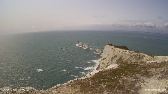 An aerial view of white chalk cliffs overlooking the ocean. There is a small building or lighthouse on top of the highest point of the cliffs. The sea below has gentle waves, with three large rocks formations rising from the water near the cliffs. The sky is partly cloudy with a horizon stretching far into the distance. The timestamp on the image reads "04/06/2025 15:00:55" and the website "isleofight.com" is visible in the bottom right corner.