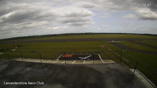 A view from a webcam overlooking the runway and grass area at Leicestershire Aero Club, with a small airplane on the taxiway in the distance. The sky is partly cloudy with patches of blue, and the date and time are displayed in the top right corner.