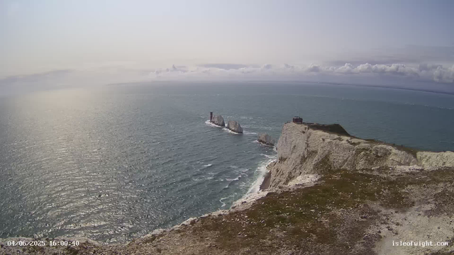 An aerial view of white chalk cliffs rising above the ocean, with a small lighthouse and a building on top of the cliffs. The sea below is shimmering under the sunlight, and there are large rocks or small islands in the water near the base of the cliffs. The sky is partly cloudy with a gentle horizon in the distance.