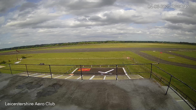 A wide view of an airfield with a grassy area and a runway in the background. In the foreground, there is a small helipad marked with a white "H" and red corners, surrounded by a metal fence. The sky is partly cloudy with patches of blue visible. The image has a timestamp in the top right corner indicating the date and time as "04-06-2025 16:00:40," and the bottom left corner has the text "Leicestershire Aero Club."