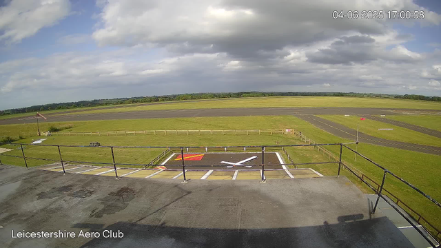 A wide view of an airfield with a grassy area, a runway in the distance, and a mostly cloudy sky overhead. In the foreground, there is a small fenced-off area with painted markings on the ground, including a large "T" and red and white markings. A flagpole with a flag is visible on the left side, and there are some small signs and barriers around the area. The timestamp indicates the date as April 6, 2025, at 17:00:58. The image is labeled "Leicestershire Aero Club" at the bottom.