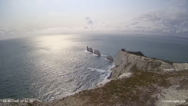 A panoramic view of a rugged coastline with tall white chalk cliffs, some topped with grassy patches, overlooking the ocean. A small building is visible on the edge of the cliff. Farther out in the sea, several large sea stacks stand in the water, with one having a lighthouse. The sky is cloudy with some sunlight reflecting off the water's surface. The timestamp in the bottom left corner indicates the image was captured on April 6, 2025, at 5:01 PM.