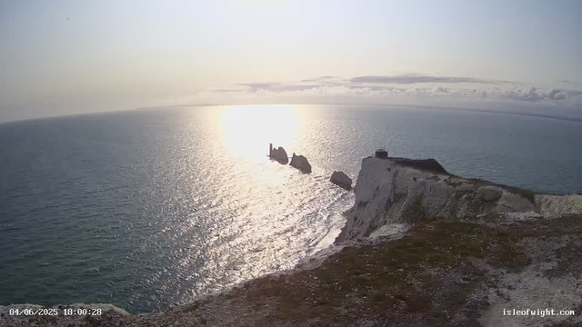 A panoramic view of a seascape at sunset or sunrise, with the sun near the horizon reflecting on the ocean. There are dramatic white chalk cliffs in the foreground, leading to a small island with a lighthouse and structures on top. The sky has a few clouds, and the overall scene is calm and scenic. The timestamp in the bottom left corner indicates the date and time as April 6, 2025, at 18:00:28. The bottom right corner has the website "isleofwight.com".