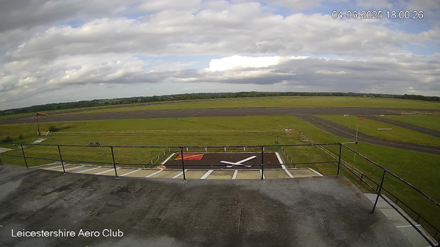 A wide view of an empty aircraft runway and grassy field at Leicestershire Aero Club on a partly cloudy day. The foreground shows a helipad with a large white "H" on a red background and a small tripod. In the background, there are open fields and a cloudy sky with patches of blue. The image has a timestamp of June 4, 2025, at 6:00 PM.