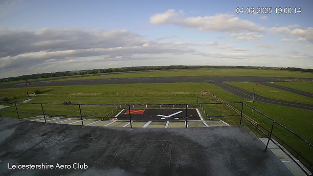 A view from a webcam at Leicestershire Aero Club showing a runway and surrounding grassy field under a partly cloudy sky. In the foreground is a large clock painted on the ground, with the clock hands pointing to approximately 4:00. The timestamp in the top right corner reads "04-06-2025 19:00:14". The scene appears calm with no aircraft or people visible.