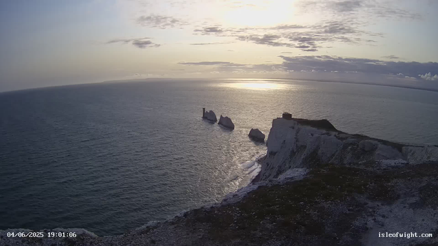 A view of white chalk cliffs overlooking the ocean at sunset. The cliffs are rugged with a small structure at the top and a pathway along the edge. In the water below, several large rock formations protrude from the sea. The sky has scattered clouds, and the sun is low on the horizon, casting a reflection on the water. The image has a timestamp in the bottom left corner and a website URL in the bottom right corner.