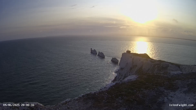 A scenic view of a coastal cliff at sunset, with white chalk formations jutting out into the sea. The sky is partly cloudy with the sun low on the horizon, casting a golden reflection on the water. There is a pathway along the edge of the cliff, and some structures are visible on top of the chalk formations. The timestamp in the bottom left corner reads "04/06/2025 20:00:16," and the bottom right corner shows the website "isleofwight.com."