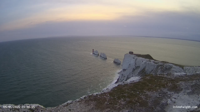A view of white chalk cliffs with a small building and a pathway along the edge, overlooking the ocean at sunset or sunrise. The sky features soft pastel colors with light clouds. The ocean waters are calm, and several large rocks are visible in the water near the cliffs. There is a timestamp in the bottom left corner showing the date and time, and a website URL in the bottom right corner.
