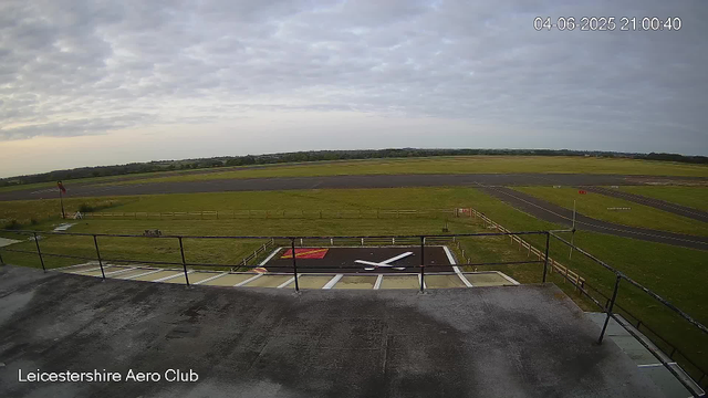 An outdoor view from a webcam at Leicestershire Aero Club showing an overcast sky with scattered clouds, a runway in the distance, and a small helipad or designated area with a white helicopter silhouette painted on the ground in the foreground. There are grassy areas, a flagpole with a red and white flag, and a wooden fence. The timestamp in the upper right corner indicates the date as June 4, 2025, and the time as 21:00:40.
