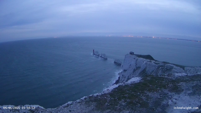 A view of white chalk cliffs and the ocean at dusk, with the sea stretching to the horizon. The sky is cloudy with shades of blue and gray. The cliffs have some structures, possibly a small tower or building, located at the top right. There are some large rocks protruding from the water near the base of the cliffs. The timestamp in the lower left corner reads "04/06/2025 21:59:53" and there is a website URL "isleofwight.com" in the lower right corner.