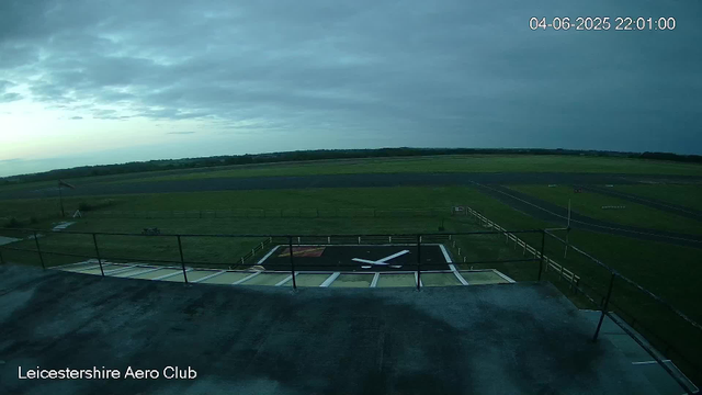 An empty aviation tarmac with a helicopter landing pad marked with a large white 'H' and an arrow pointing forward. A black railing surrounds the helicopter pad, which is situated on a raised platform. Beyond the platform, there is a vast open green field with a few small structures and pathways. The sky is mostly cloudy with some patches of lighter sky near the horizon. The top right corner displays a timestamp: 04-06-2025 22:01:00. The bottom left corner has the text "Leicestershire Aero Club."