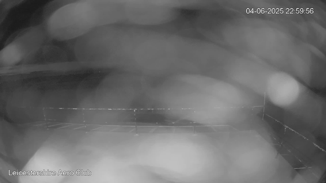 A black-and-white image from a webcam shows a blurry, indistinct scene with some structures that appear to be part of an outdoor area. There is a timestamp in the top right corner displaying "04-06-2025 22:59:56," and in the bottom left corner, the text reads "Leicestershire Aero Club." The overall image is hazy and lacks clear details.