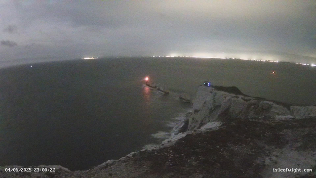 A nighttime view of a coastal scene with a rocky cliff in the foreground. The ocean extends into the distance, with a few navigational lights visible on the water. The sky is cloudy with some illumination near the horizon, likely from distant city lights. There is a timestamp in the lower left corner reading "04/06/2025 23:00:22" and a website address "isleofwight.com" in the lower right corner.