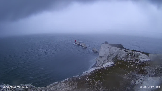 A view of white chalk cliffs stretching into the ocean, with three small islands or rocks near the coast. A red and white lighthouse stands on one of the islands. The sky is overcast with dark clouds, and the water appears calm with gentle ripples. The scene looks misty and cool.
