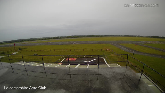 An empty airport tarmac with a control tower and runway in the distance under a cloudy sky. The image is from a webcam at Leicesterhire Aero Club, with a timestamp of May 6, 2025, at 05:00:51. In the foreground, there is a helipad with a white outline and a large white 'H' in the center. The helipad is surrounded by a safety fence, and the scene appears to be taken from an elevated position overlooking the airfield.