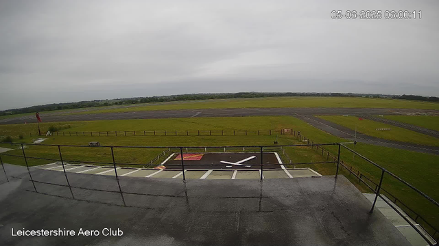 A view of an airfield from a webcam at Leicester Aeroclub, showing a cloudy sky, a grassy area with runways in the background, and a rooftop helipad in the foreground. The scene appears calm and overcast, with no aircraft or people visible. The timestamp indicates the photo was taken on June 5, 2025, at 6:00 AM.