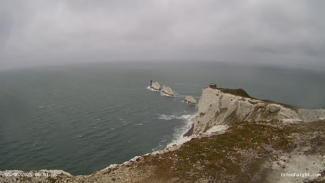 A view of steep, white chalk cliffs extending into the ocean under a cloudy sky. There is a small building on top of the closest cliff, and further along the coast, a lighthouse stands on a rock formation surrounded by water. The sea appears choppy with waves, and the atmosphere is overcast and moody. The image is timestamped "05/06/2025 06:01:06" and includes a website URL "isleofwight.com" in the bottom right corner.