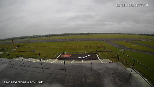 A view from a webcam at Leicestershire Aero Club shows a cloudy sky overhead, green grass fields, and a small helicopter landing pad in the foreground with a large clock painted on it. There is a windsock to the left and a fenced area surrounding the landing zone. The image also includes a timestamp in the top right corner showing the date and time.