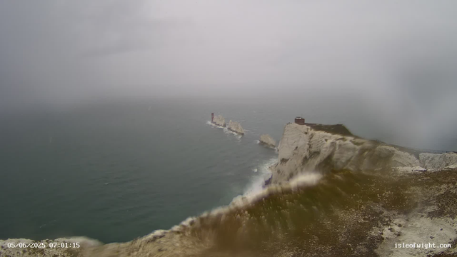 A view of white chalk cliffs with a lighthouse on top, overlooking the ocean under an overcast sky. Waves crash against the base of the cliffs, and a distant rocky formation extends into the water. The scene is gloomy with gray clouds and misty atmosphere. The timestamp in the lower left corner reads "05/06/2025 07:01:15," and there is a website URL "isleofwight.com" in the lower right corner.