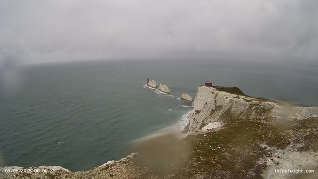 A view of white chalk cliffs and rock formations extending into the sea under a cloudy, gray sky. At the top of the cliffs, there is a small building or lighthouse, and further out in the water, a red lighthouse stands on a separate rock formation. The scene appears to be captured from a webcam, with a timestamp in the lower left corner.