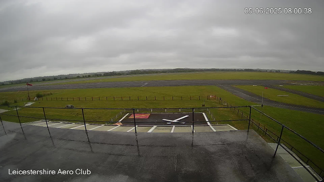 A view from a webcam overlooking an airfield on a cloudy day. In the foreground, there is a helipad with a large white "H" surrounded by a red square. A black and white clock is painted on the tarmac, and a small flagpole with a red windsock is visible on the left side. The grassy area beyond is fenced, with a picnic table and some bushes. The runway stretches into the distance, with a few markings and signs, and rolling green fields extend toward the horizon under a gray, overcast sky. The timestamp in the top right corner reads "05-06-2025 08:00:38," and the bottom left corner displays "Leicestershire Aero Club."
