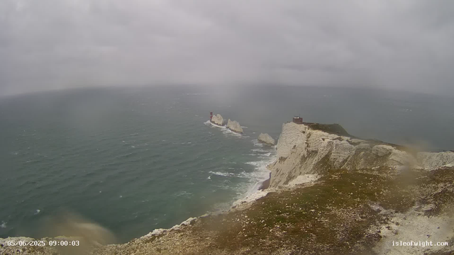 A view from a webcam showing a rugged coastline with white chalk cliffs and a small building on top. In the distance, there are smaller rock formations in the sea, one of which has a red lighthouse on it. The sky is overcast with thick gray clouds, and the sea appears choppy with small whitecaps. The timestamp in the lower left corner indicates the date and time as May 6, 2025, at 9:00 AM. The website "isleofwight.com" is visible in the lower right corner.