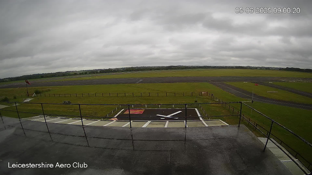 An overcast sky above an airfield with a runway stretching into the distance. In the foreground, there is a helipad with a large white and black H symbol and a red square marking, surrounded by a fence. To the left, a windsock is attached to a pole. The bottom left corner of the image has the text "Leicestershire Aero Club."