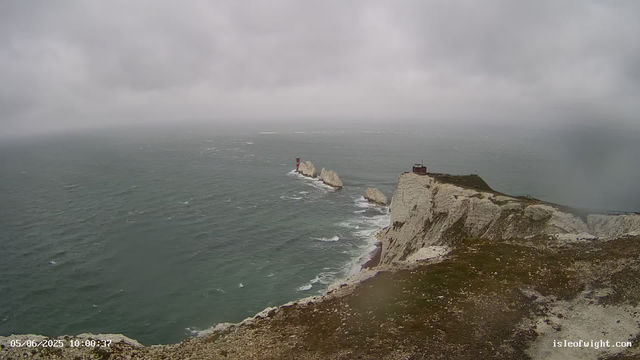 A view of high white chalk cliffs on a rugged coastline under a cloudy sky. The sea below is choppy with waves, and several large rocks and a small lighthouse are visible near the base of the cliffs. There is a small building or structure at the top of the cliff. The timestamp on the image reads "05/06/2025 10:00:37" and the website "isleofwight.com" is displayed in the bottom right corner.