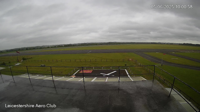 A wide view of an overcast day at Leicestershire Aero Club, showing a runway in the distance, a grassy field in the middle, and a parking area with a large clock on the ground in the foreground. The image includes a date and time stamp in the upper right corner and is taken from a webcam mounted on a balcony or terrace. The sky is cloudy and there are no visible aircraft or vehicles in motion.