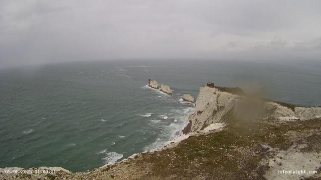 A view of white chalk cliffs rising from the sea under a cloudy sky. There is a small, dark-colored building atop the cliffs on the right and several white rock stacks with a red lighthouse on one of them in the ocean to the left. The water appears turbulent with whitecaps, and the coastline extends into the distance. The image timestamp shows "05/06/2025 11:00:21" and the website "isleofwight.com" is visible in the bottom right corner.