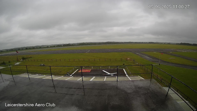 An overcast sky covers the Leicestershire Aero Club's airfield. The view includes a grassy area with a windsock on a pole, some picnic tables, and a small fenced section with runway markings on the ground. The runway and taxiways extend into the distance, with open fields and a tree line in the background. The timestamp indicates the photo was taken on June 5, 2025, at 11:00 AM.