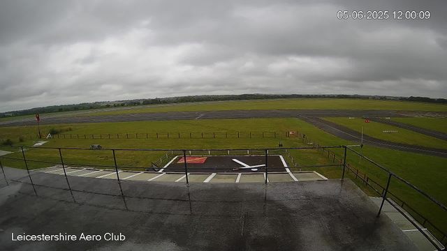 A view from a webcam at Leicestershire Aero Club showing an overcast sky with thick gray clouds. The image captures the runway and surrounding grassy area, with a small helipad in the foreground marked with a red square and a white helicopter symbol. A black safety barrier lines the edge of the platform, and in the distance, there are open green fields and a distant tree line. The timestamp in the upper right corner indicates the date as June 5, 2025, and the time as 12:00:09.