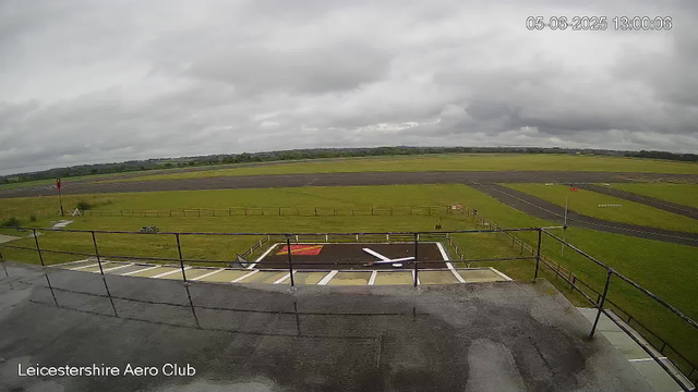 A view of an airfield under a cloudy sky. In the foreground, there is a platform with a black railing and a helicopter landing pad marked with a white circle and a white arrow pointing right. To the left, there is a small picnic table. Beyond the platform, there are well-maintained grassy areas, paved runways, and taxiways stretching into the distance. The timestamp shows "05-06-2025 13:00:06," and the bottom left corner has the label "Leicestershire Aero Club."