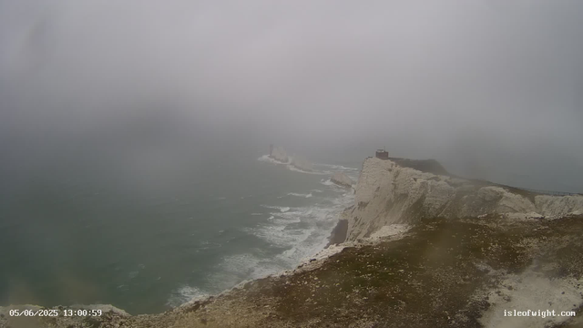 A view of white chalk cliffs along the coastline on a foggy day. The cliffs descend sharply into the sea, with waves crashing at their base. There is a small building or lighthouse near the edge of the cliff, and a pathway or railing runs along the cliff top. The overall scene is misty, obscuring the horizon and distant parts of the coastline. A timestamp in the bottom left indicates the date and time as 05/06/2025 13:00:59.