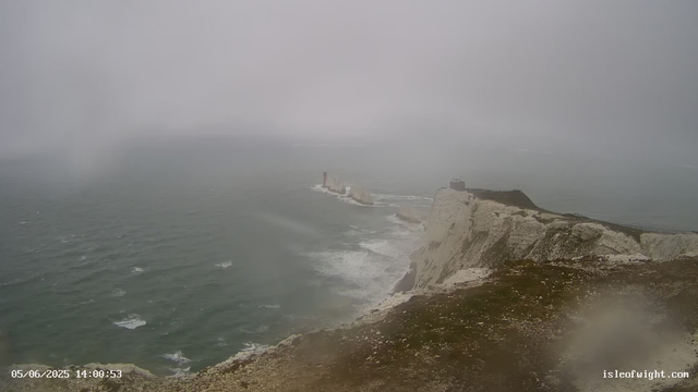 A foggy daytime scene of white chalk cliffs along the coast with a small, dark building near the edge. The cliffs rise sharply from the ocean below, where waves are crashing against the rocks. A tall lighthouse is visible farther along the cliffs, and a narrow pathway runs along the top of the cliffs. The scene appears to be captured from a webcam, with a timestamp of 05/06/2025 14:00:53 and the website isleofwight.com in the lower right corner.
