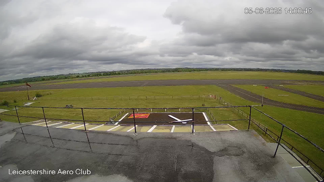 A view from a webcam at Leicestershire Aero Club shows a cloudy sky over an open grassy area with a runway in the distance. In the foreground, there is a parking area with white lines and a red and white marked helipad. The tarmac appears wet, possibly due to recent rain. A small bench and a windsock are visible towards the left side of the image. The timestamp indicates it is 14:00 on June 5, 2025.