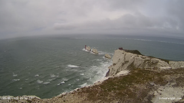 A wide view of a rugged coastal landscape with white chalk cliffs and large rock formations in the ocean. A small building or lighthouse is situated on the edge of the cliff on the right, overlooking the sea. The sky is overcast with gray clouds, and the ocean appears choppy with waves. In the distance, a faint white line of a distant boat's wake can be seen across the sea. The timestamp on the image indicates it was taken on June 5, 2025, at 15:00.