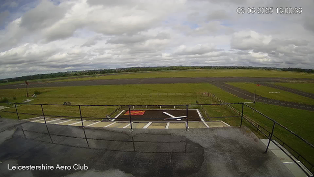An outdoor view from a webcam at Leicesterhire Aero Club shows a small aircraft landing pad with a white tail number on a black background. There is a parking area with marked spaces and a black ramp with a white arrow pointing toward the runway. The scene includes a fence, a grassy field, a wind sock, and a flat, open landscape extending into the distance under a cloudy sky. The timestamp in the top right corner reads "05-06-2025 15:00:36."