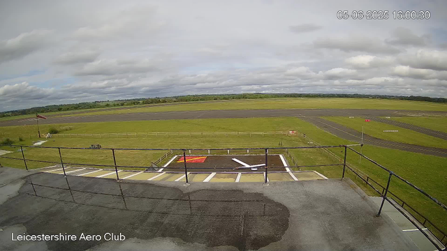 A view from a webcam showing an open-air area within Leicestershire Aero Club. In the foreground, there is a concrete platform with black metal railings, some painted markings on the ground, and a small windsock in the grass to the left. Beyond that, there are grassy fields, taxiways, and runways stretching into the distance. The sky is mostly cloudy with patches of blue, and the overall scene is calm and spacious, with no aircraft or people visible. The date and time displayed are 05-06-2025 16:00:30.