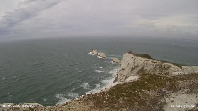 A view of the white chalk cliffs of Étretat on the coast of Normandy, France. The image shows the cliffs extending into the sea with rock formations and a lighthouse on the rocks in the distance. The sky is overcast with gray clouds, and the ocean appears choppy. There is a small building or structure on the edge of the cliffs. The timestamp indicates the photo was taken on June 5, 2025, at 16:00:56.