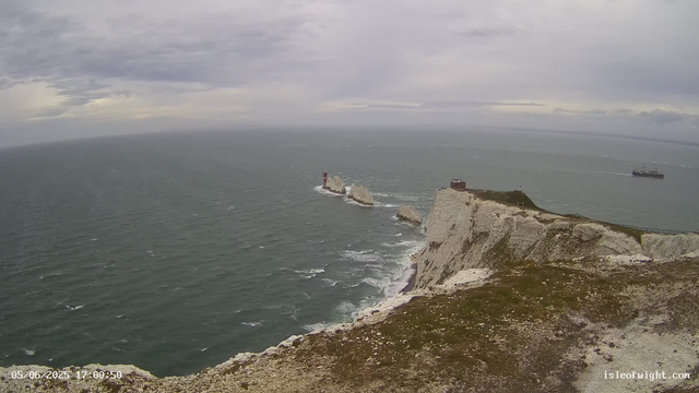 A view from a webcam shows white chalk cliffs extending into the ocean, with a lighthouse on one of the smaller offshore rocks. The sea appears choppy with small waves, and there is a ship sailing in the distance. The sky is cloudy and overcast. The timestamp on the image indicates it was taken on May 6, 2025, at 17:00:50. The website "isleofwight.com" is visible in the bottom right corner.