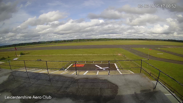 A view of an outdoor area at Leicesterhire Aero Club under partly cloudy skies. The foreground shows a concrete platform with a black railing, and a designated helicopter landing spot marked with a large white arrow and a red and yellow square. Beyond the platform, there is a grassy field with a fence, a small sign, and a windsock. In the distance, there are taxiways, runways, and open fields extending to the horizon. The timestamp in the top right corner indicates the date as June 5, 2025, and the time as 17:00:52.