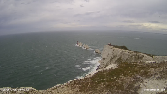 A view of high white chalk cliffs overlooking the ocean on a cloudy day. There is a small structure on top of the nearest cliff, and a lighthouse with a red top situated on a rock formation offshore. The sea appears slightly choppy, and the sky is mostly overcast with some patches of lighter clouds. A narrow path runs along the edge of the cliff. The timestamp indicates the photo was taken on June 5, 2025, at 6:00 PM.
