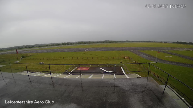 A view from a webcam at Leicestershire Aero Club showing a wet runway under a cloudy, overcast sky with no visible aircraft or people. The foreground features a platform with railings and a red and white markings area, while the background displays grass fields and taxiways stretching into the distance. The timestamp reads 05-06-2025 18:00:52.