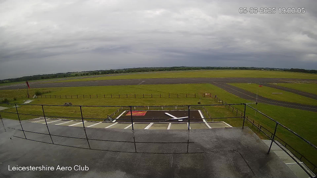 A view of an airfield under a cloudy sky, with a takeoff or landing area in the foreground. There is a small parking area with white lines, a landing pad marked with red and white, and black passenger tarmac with white markings. The airfield is surrounded by green fields and a distant tree line. The timestamp in the top right corner shows "05-06-2025 19:00:05," and the bottom left corner indicates it is located at the "Leicestershire Aero Club."