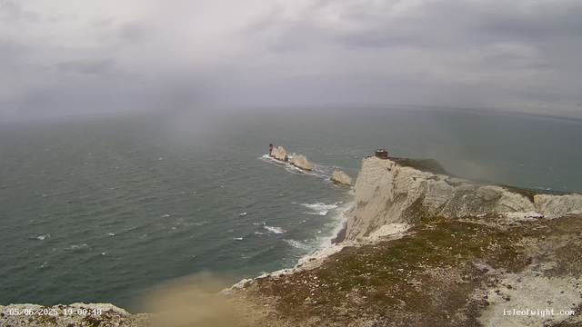 A view of white chalk cliffs extending into the ocean under a cloudy sky. The cliffs have green patches at the top, and there are rocks and a lighthouse visible on one of the distant rock formations in the water. The scene appears to be taken from a webcam, with a timestamp of 05/06/2023 at 19:00:48.