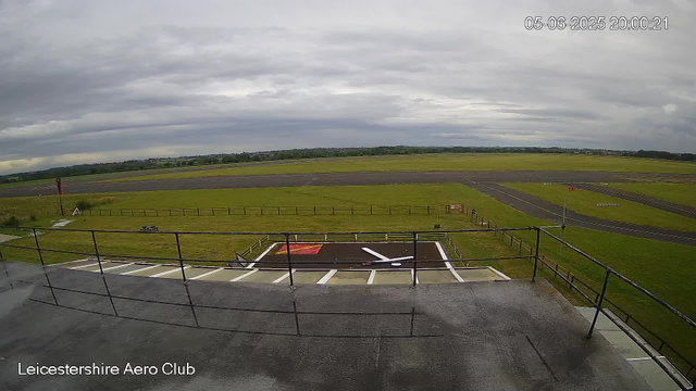 A view from a webcam showing the runway and surrounding grassy area at Leicestershire Aero Club. The sky is mostly cloudy with some breaks in the clouds. There are railings in the foreground, and a marked helipad with a letter "H" and white lines. The timestamp in the top right corner indicates it was taken on May 6, 2025, at 20:00:21.